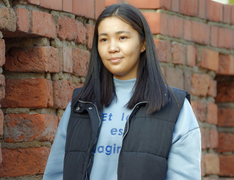 27 - Person standing in front of a rustic brick wall, wearing a light blue top and a black puffer vest
